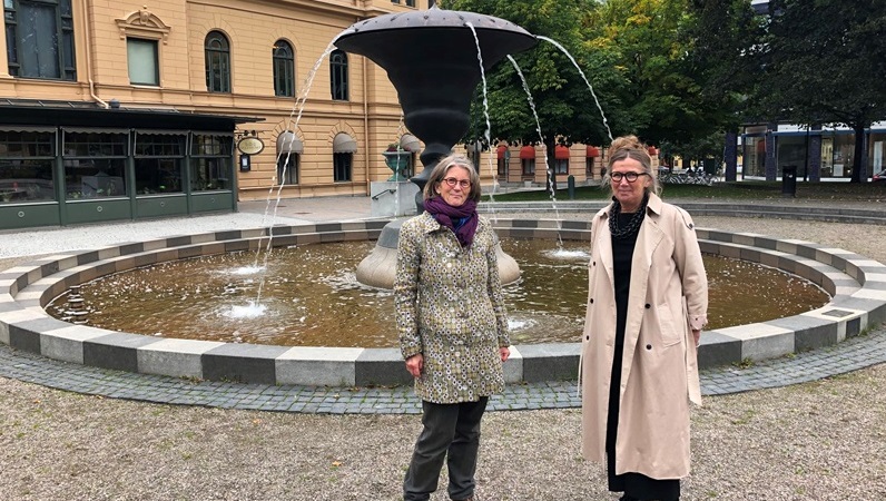 Två leende kvinnor står framför en hög, fontänskulptur i en stadspark.Kvinnorna är i övre medelåldern. Den ena har grått hår i p