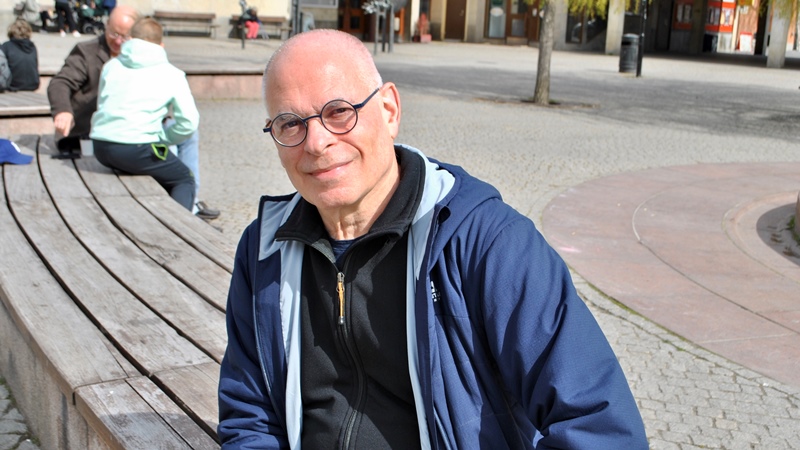 Leende äldre man med glasögon och blå sportjacka, sitter på en bänk