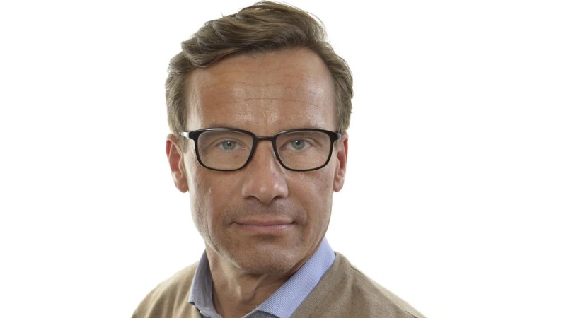 Porträtt på moderaten Ulf Kristersson. Han har ljusbrunt hår, gröna ögon bakom glasögon med mörka bågar och är klädd i ljusblå skjorta under beige tröja.