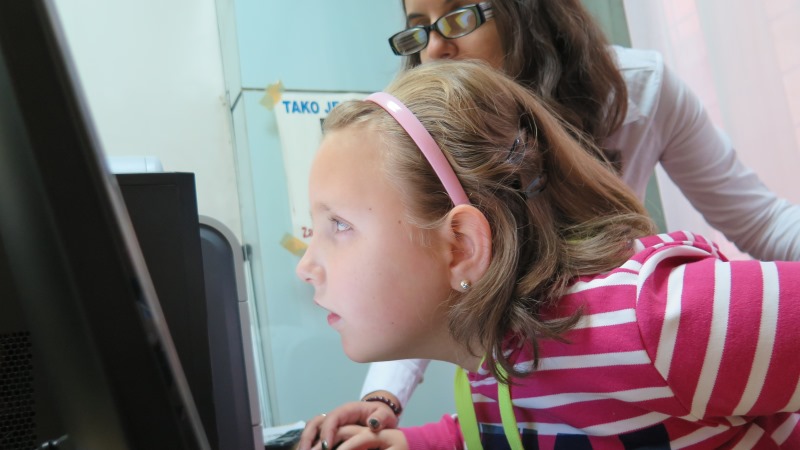 Flicka med halvlångt blont hår och rödvitrandig tröja med ansiktet nära en datorskärm. I bakgrunden en vuxen som håller hennes hand och tittar över hennes axel.