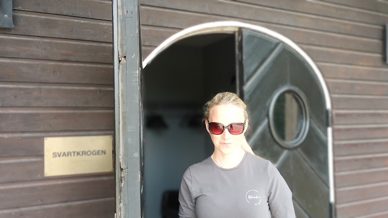 En blond kvinna i tonade glasögon står i en välvd port. På en skylt på sidan står det Svartkrogen.
