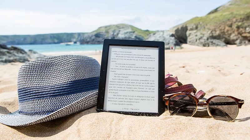 På en sandstrand ligger en e-boksläsare mellan ett par mörka glasögon och en blå halmhatt. I bakgrunden suddiga klippor och vatten.
