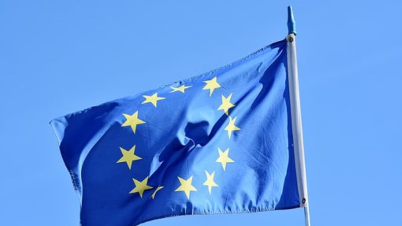 Närbild på EU.s blå flagga, med en stor cirkel av små gula stjärnor, som vajar i vinden mot en ljusare blå himmel.