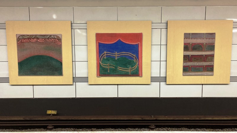 Tre tavlor med olika motiv och varmgula passpartouer av konstnären Lisa Lundgren hänger på en plattformsvägg över spåret.