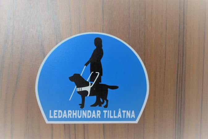 Ett blått klistermärke med den vita texten "Ledarhundar tillåtna" och en stiliserad bild på ett ledarhundsekipage. Klistermärket sitter på en dörr i träpanel.