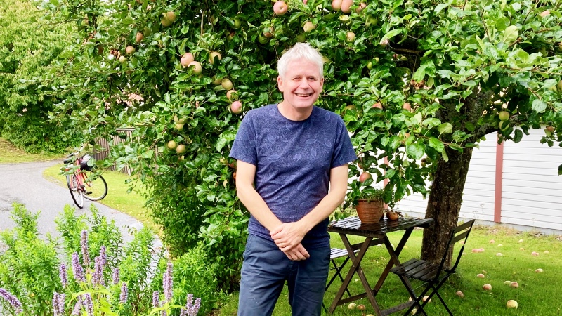 Leende gråhårig man i blå T-tröja står under äppelträd ( sommar)