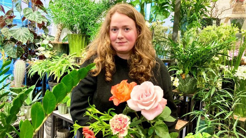 Madeleine Hobring, en ung kvinna med långt mörkblont hår omgiven av gröna blad och med ljusa rosor i famnen.