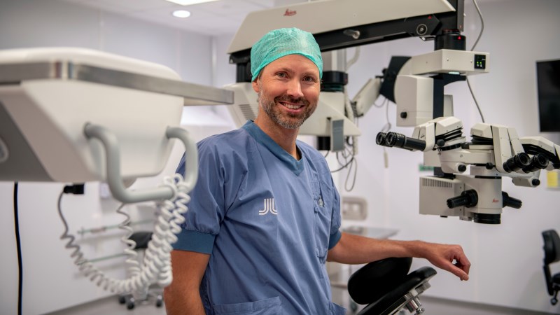 Vit medelålders man med skäggstubb, i ljusblå läkarkläder och mintgrön operationsmössa. Står i operationssal med stort hängande mikroskop.