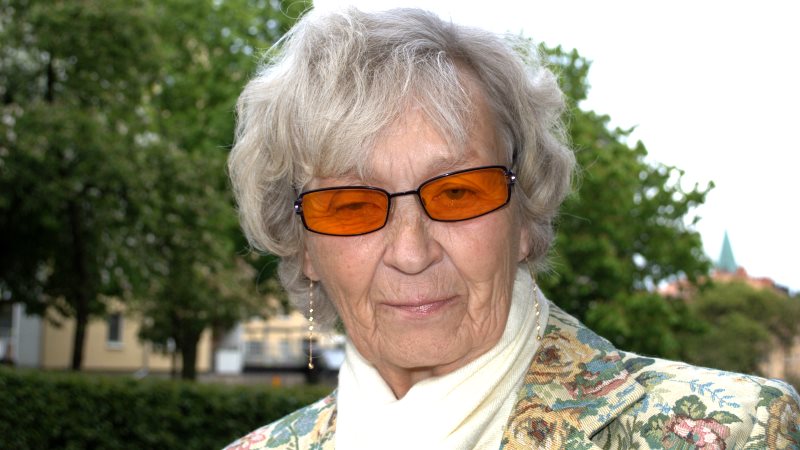 Maud Tillgren har grått, ganska kort hår, orangea glasögon, mönstrad kavaj och vit scarf.