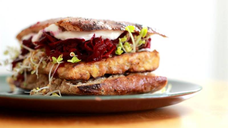 Bild på hamburgare med tunnt mörkt surdegsbröd, grönsaker och vad som liknar en stor ostbit i mitten.