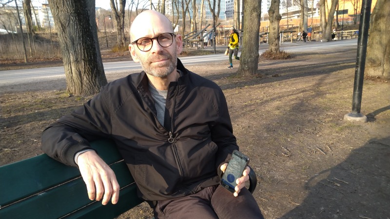 En skäggig man med glasögon och kalt huvud sitter på en parkbänk med en mobiltelefon i handen där man skymtar SOS Alarm-appen.
