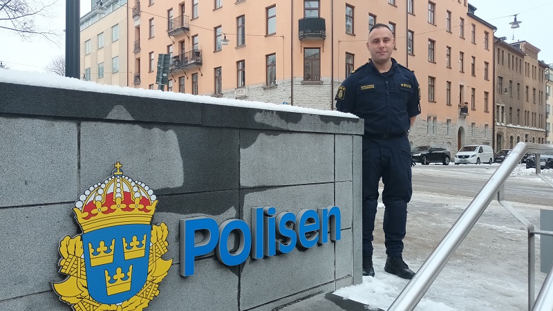 Daniel Momeni, en kortklippt mörkhårig vit man i polisuniform står i stadsmiljö intill en stenmur med ordet Polisen i blått och myndighetens emblem i gult, blått och lite rött.