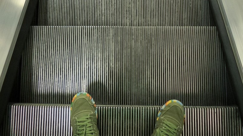 Tre steg i en rulltrappa sedda uppifrån. Ett par gröna skor sticker fram i bildens nederkant.