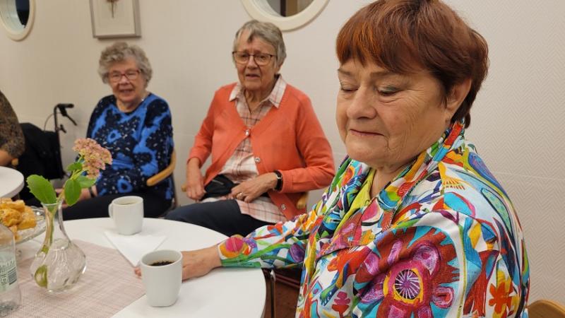 Tre äldre vita kvinnor i bild, hon som är i förgrunden har rödlätt hår, grön ögonskugga och en kavaj i flera färger.