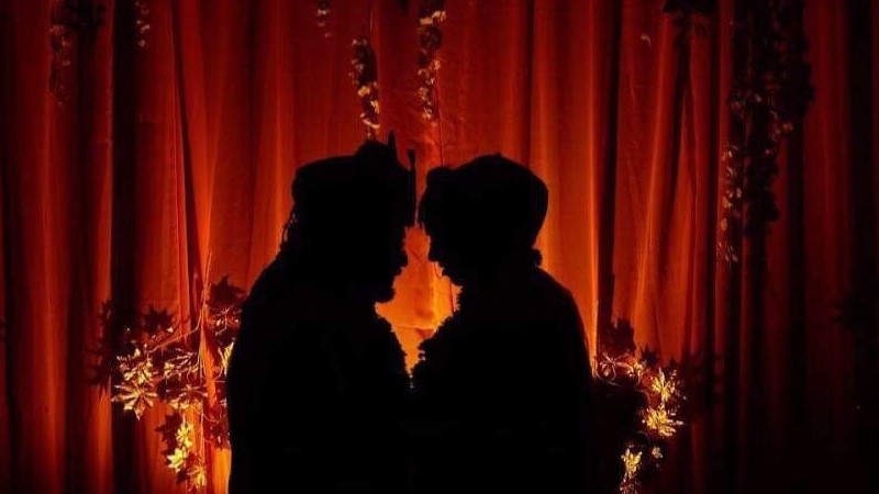 Silhuetterna av två personer som står med ansiktena lutade mot varandra och håller om varandra syns mot en bakgrund av en belyst röd ridå.