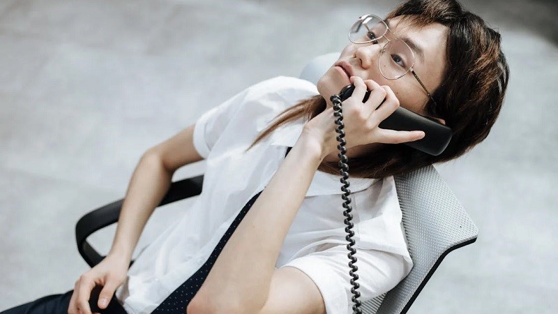 En person med androgynt utseende i glasögon och mörkt hår sitter med en sladdbunden telefonlur mot örat tillbakalutad i en kontorsstol.