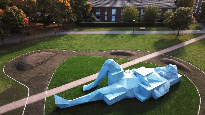 Enorm ljusblå betongskulptur av liggande man på gräsmatta i park