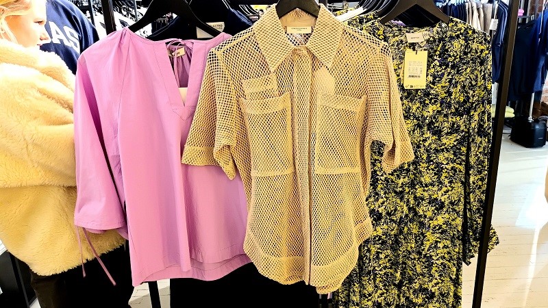 Tre plagg hänger i förgrunden i en butik full med klädställningar. En rosa topp med vida ärmar, en kortärmad beige skjorta av nät och en lång klänning med marmormönster blått gult och svart.