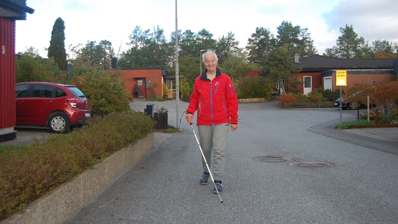 Äldre man i röd jacka och med vit käpp går på gata i villaområde