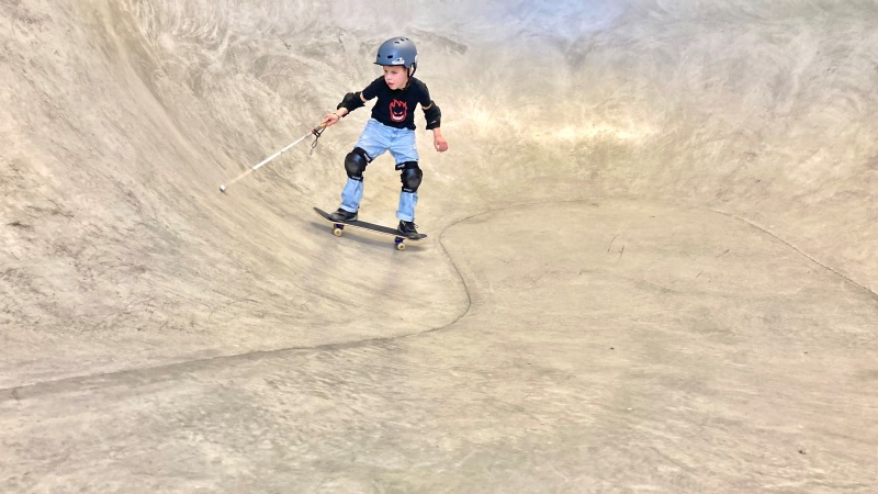 En pojke åker skateboard med sin vita käpp i en cementbeklädd ramp ( grop). Har p sig hjälm, knäskydd och blå jeans.